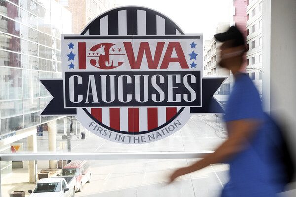 A blurry person walks past a sign on a window reading "Iowa Caucuses, first in the nation" with an illustration of an elephant and a donkey inside the "o" in Iowa.