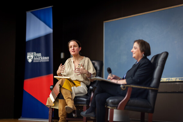 Sophia Rosenfeld and Beth Wenger sit on chairs on a wooden stage in front of a black board and a sign reading Penn Arts &amp; Sciences, as Rosenfeld addresses the audience.