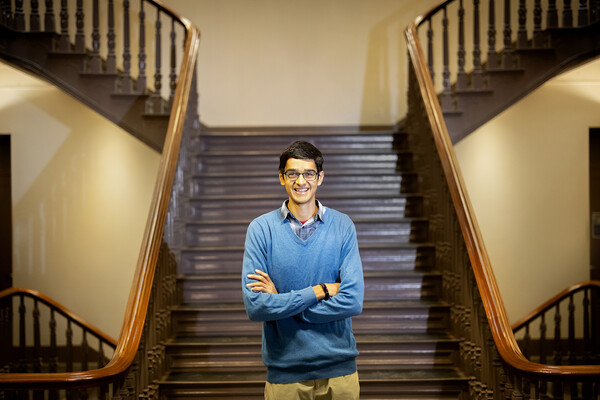 Wearing glasses and a blue sweater, Om Manghani stands with crossed arms on a staircase