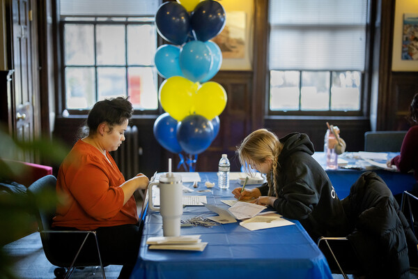 Two people face each other at a table, filling out paperwork, with a tower of blue and yellow balloons in the background