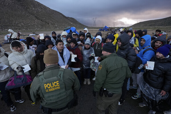 A group of migrants along the Mexico-California border show their identification to U.S. Border Patrol agents, with brown mountains in the background and the sun about to rise, giving a spot of light in an overcast sky.