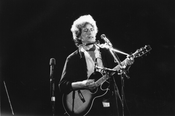 Bob Dylan on stage playing his guitar with his harmonica on a holder near his face.