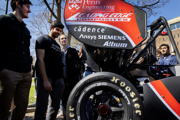 A group of students is gathered around a Formula-style racecar at an event.