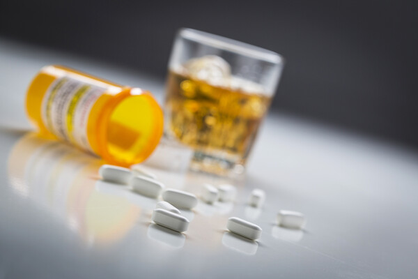 An open bottle of prescription pills spilling onto a table with a tumbler of whiskey.