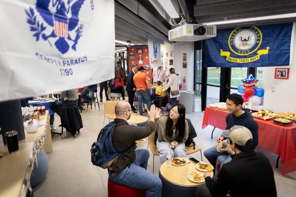 A room full of people eating lunch with flags from all the Armed Services hanging from the rafters. 