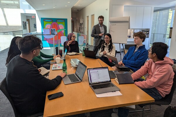 A group of students sits around a rectangular table in a discussion. A man stands next to a pad of paper on an easel preparing to take notes.