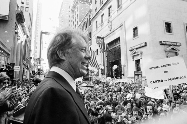 In a black-and-white photo, Jimmy Carter stands close to the camera in front of a crowd. He is seen in profile and is smiling.