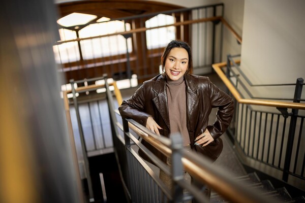 A woman in a brown jacket and brown shirt stands in a stairwell smiling at the camera.