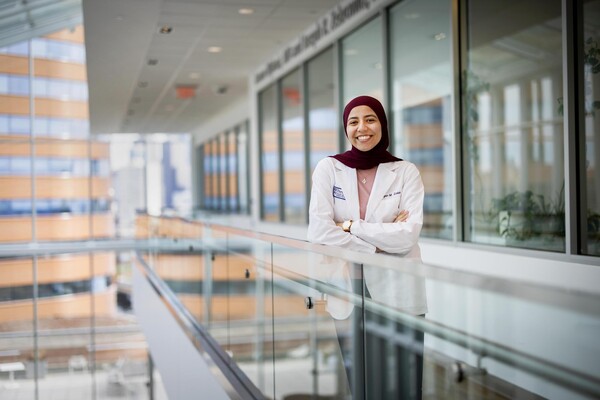 A smiling woman stands facing the camera with her arms crossed. She is standing in front of a glass-fronted row of offices with a glass railing in front of her. She is wearing a white coat and maroon head covering.