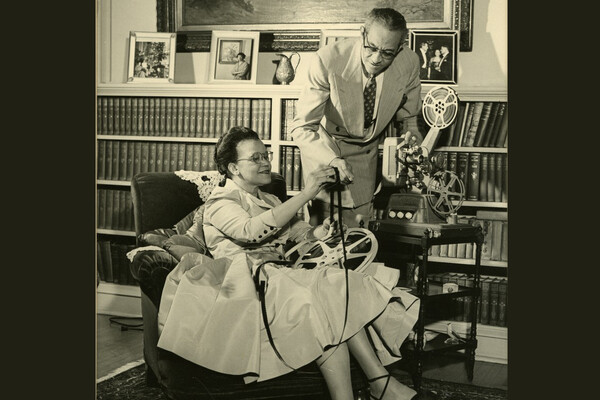 Sadie and Raymond Alexander with a film projector in a room with books on bookshelves and framed photos behind them. 