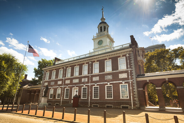 A brick building with white windows is seen against a blue sky. An American flag flies to the left, and a clock sits in the cupola at the center. 