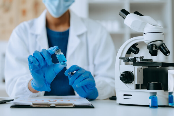 A researcher holding a vaccine vial in a lab next to a microscope.