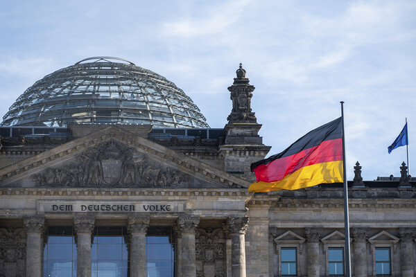 The dome of a building is visible on the left. To the right, a striped black, red and yellow flag is flying on a pole. An inscription on the building reads: “Dem Deutschen Volke,” or “To the German People.”