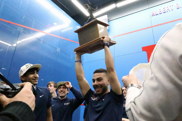 A member of Penn’s squash team holds a squash trophy over their head with other teammates.