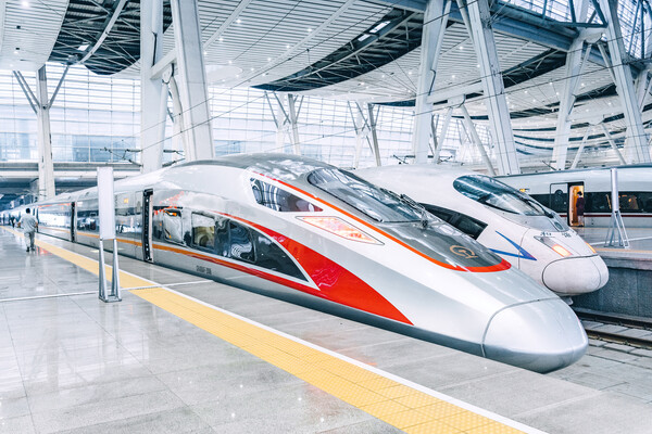 Two bullet trains sit side-by-side in a silver-and-white train station. The train closest to the camera has red markings on the side and top.