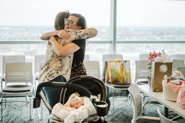 Two people embrace at a uterine transplant meetup lunch with a baby in a stroller and gifts on a conference table.