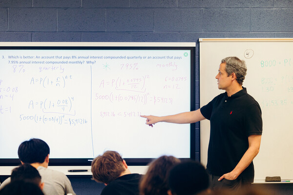 A teacher pointing ot a whiteboard in a high school classroom.