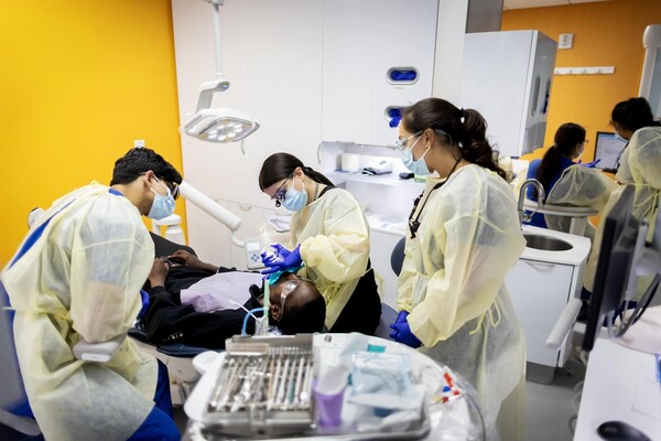 3 Penn Dental Med students at the clinic, one is cleaning a patient’s teeth.