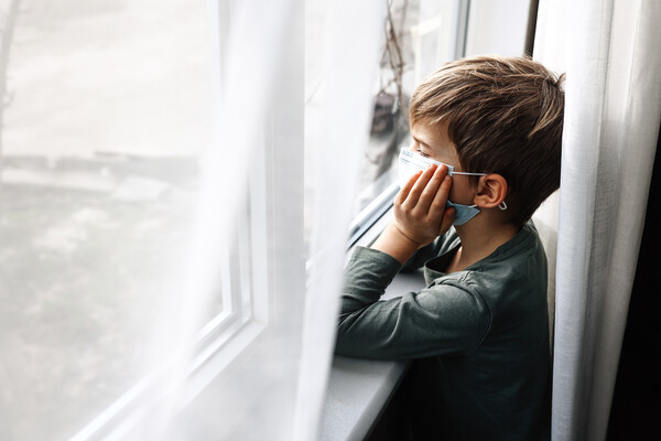 A young child at a window wearing a mask due to COVID.