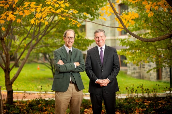 Michael Ostap, left, with David Meaney outdoors in front of a limestone building with autumn trees, wearing suits and ties.