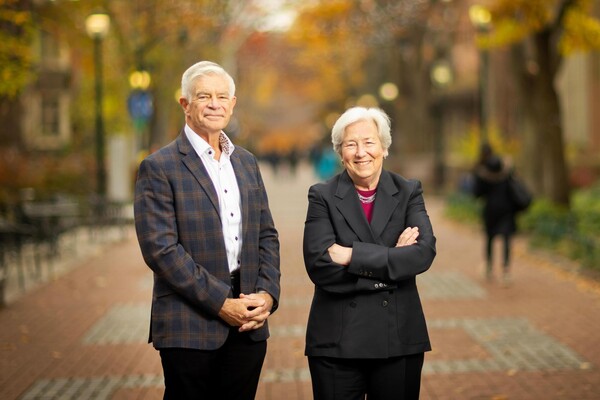 Two people standing on a brick pathway lined with trees in autumn, wearing formal business attire.