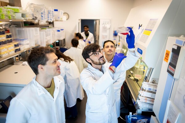 Marco Ruella in the Ruella Lab, collaborating with two lab technicians. Ruella is wearing a white lab coat and holding up a chemical sample. Two of his colleagues are observing the sample.