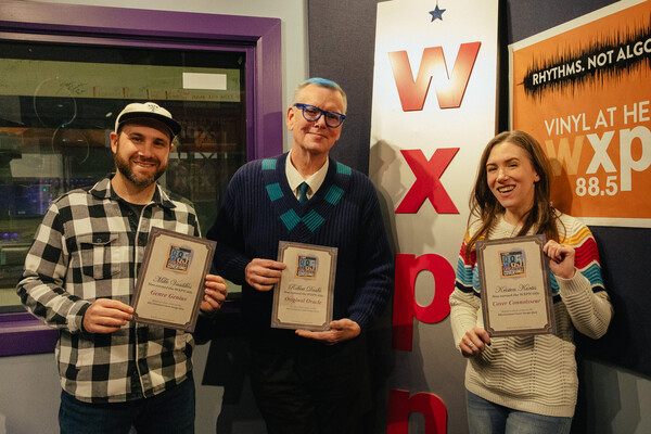  Three people standing in a WXPN radio studio holding framed certificates, with a large WXPN sign and station branding visible in the background.