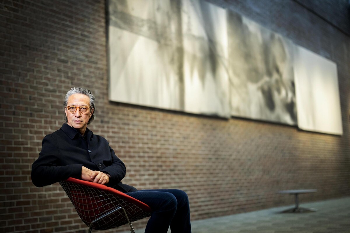 Makoto Fujimura rests his arms on a chair in front of a triptych of paintings.