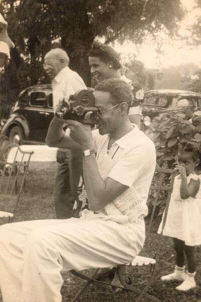 Raymond Alexander holding a movie camera while sitting in a chair outside with cars and flowering bushes behind him. 